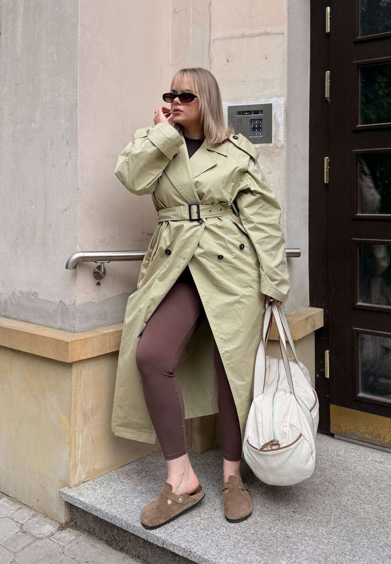 Femme portant un trench beige, des leggings marron et des chaussures marron sans lacets, tenant un sac blanc, debout à un coin de bâtiment avec une porte.