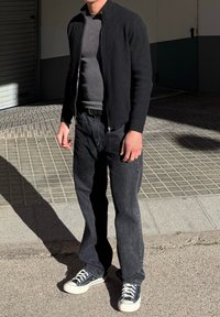 Άνδρας με μαύρο σακάκι με φερμουάρ, γκρι πουκάμισο, σκούρο τζιν και μαύρα-άσπρα αθλητικά παπούτσια, όρθιος σε πλακόστρωτο πεζοδρόμιο με σκιά στο έδαφος.