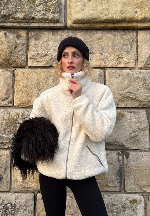 Chaqueta blanca y peluda con cremallera, cuello alto, bolsillos laterales y accesorio de pelo sintético negro texturizado, combinada con leggings negros frente a una pared de piedra.