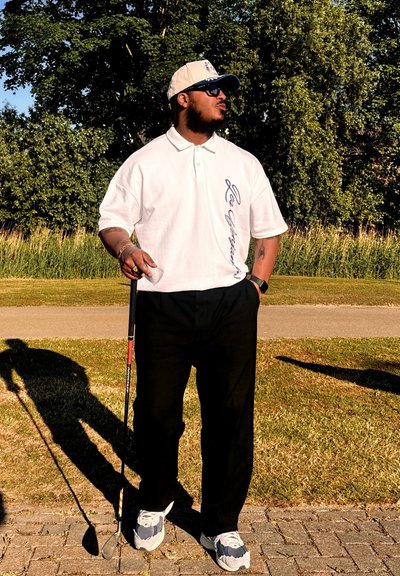 Camisa polo blanca con texto bordado, pantalones negros y zapatillas. Lleva una gorra beige y gafas de sol, sosteniendo un palo de golf.