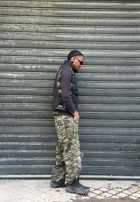 Sort puffervest, langærmet skjorte og camouflage posede bukser. Sorte chunky støvler med teksturerede såler. Mørk baggrund med korrugeret mønster.