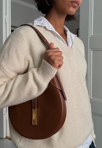 Braune Lederhandtasche in runder Form mit goldenen Akzenten und Stickdetails, gehalten von einer Person, die einen cremefarbenen Pullover und ein weißes Hemd trägt.