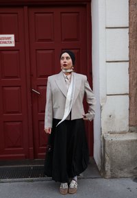 Blazer beige ajusté sur une chemise claire, associé à une jupe noire et des baskets beiges. Comprend une �écharpe blanche avec des rayures noires.