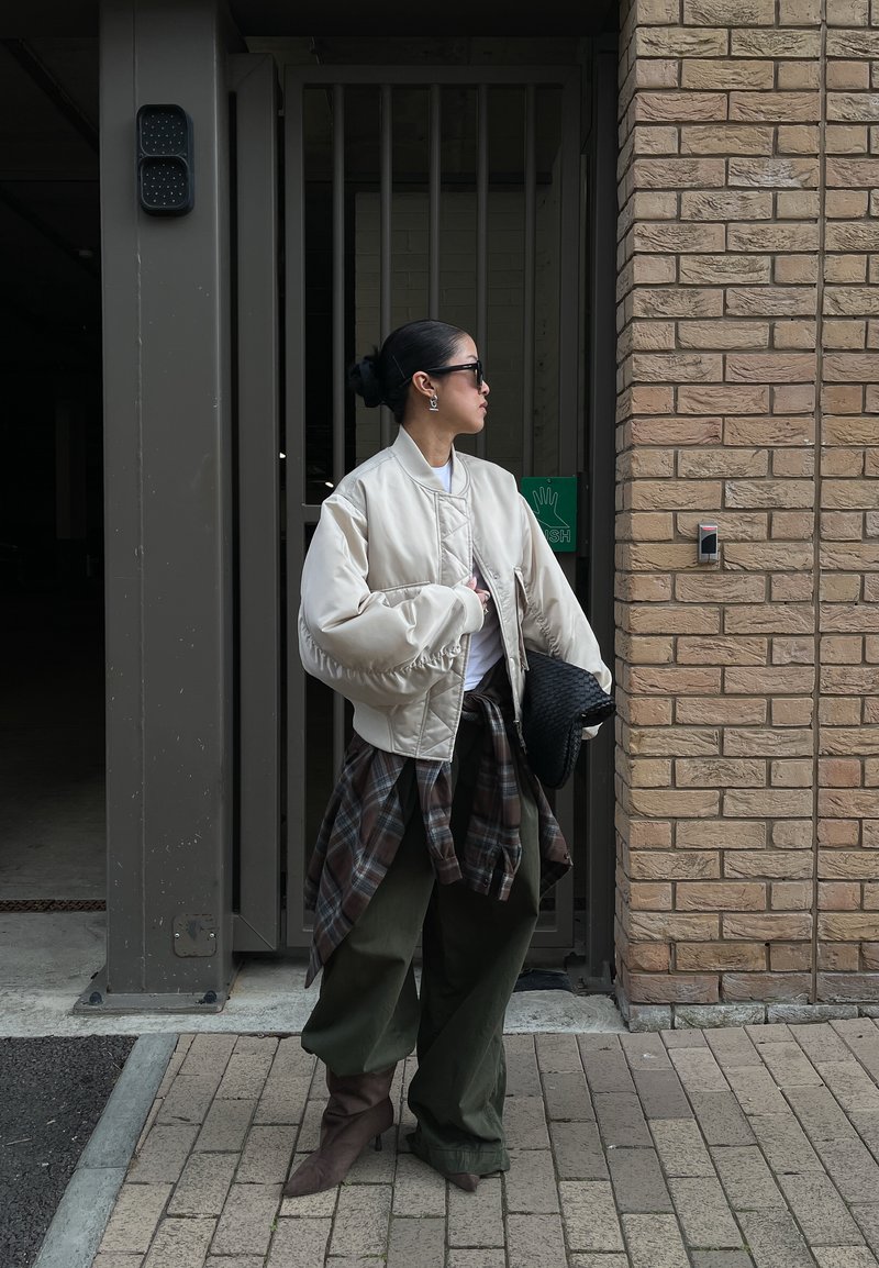 Frau in übergroßer beiger Jacke, kariertem Hemd, das an der Taille gebunden ist, olivgrünen weiten Hosen, braunen Stiefeln, Sonnenbrille und schwarzer Clutch steht nahe einer Ziegelmauer.