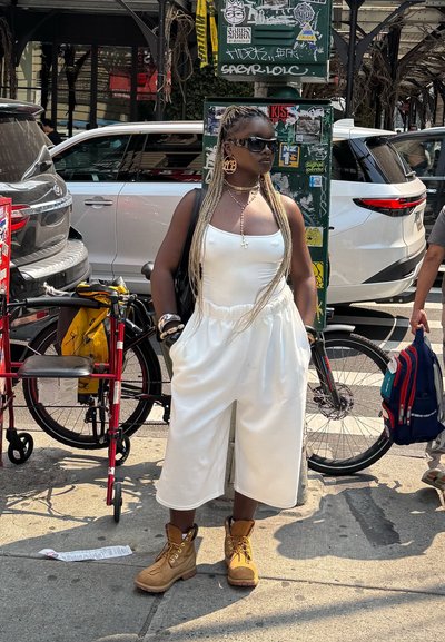 Mono blanco con tirantes de espagueti y piernas anchas, combinado con botas beige. Cabello largo trenzado y grandes aretes de aro. Bicicleta y silla de ruedas al fondo.