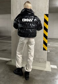 Černá lesklá péřová bunda s logem DKNY na zadní straně, spárovaná se stříbrnými lesklými kalhotami a černými kotníkovými botami v betonovém prostředí.