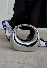 Escultura em cromado brilhante com uma forma suave e arredondada e um centro aberto. Posicionada sobre um piso de concreto perto de sapatos atléticos pretos e prateados.