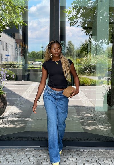 Una persona con una camiseta negra ajustada y vaqueros de pierna ancha azules sostiene una caja marrón mientras camina por unas puertas de cristal rodeadas de un jardín verde.