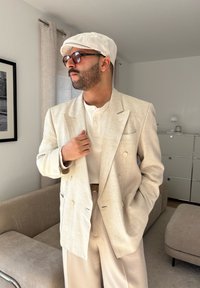 Blazer duplo em linho bege claro, camisa henley branca com texturas, calças bege, boné cinza e óculos de sol castanhos. Design simples e bem ajustado.