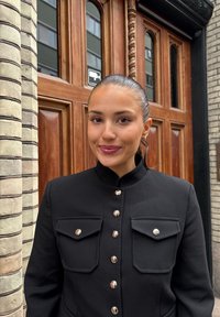 Sort jakke med høy krage, trykknapper og to frontlommer. Glatt stoff med skreddersydd passform. Bakgrunnen viser tredører.