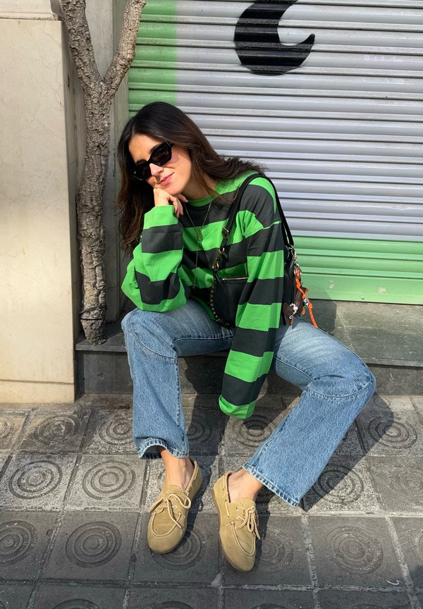 Maglietta a maniche lunghe verde e nera a righe, jeans a campana azzurri chiari, scarpe slip-on beige con lacci e una piccola borsa nera.