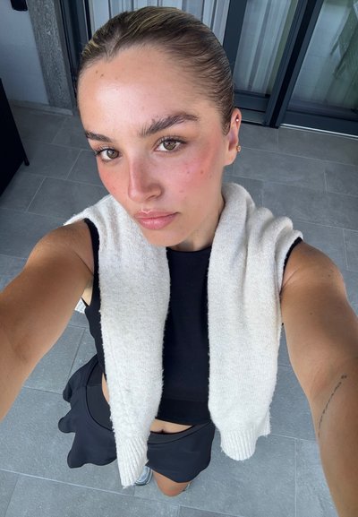 Top negro sin mangas, chal blanco de punto, falda negra y zapatos plateados. Piel suave con rubor rosa pálido, ojos marrones y un peinado de moño elegante.