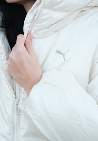 Veste blanche matelassée avec capuche, en tissu brillant, fermeture éclair à l'avant et petit logo Puma gris sur la poitrine. Manches texturées visibles.