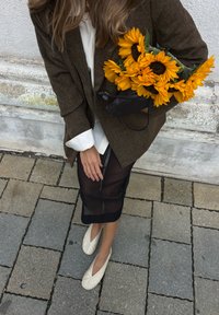 Brązowa, teksturowana marynarka nałożona na białą koszulę, w zestawieniu z czarną, przezroczystą spódnicą. Trzymając bukiet jasnożółtych słoneczników, ma na sobie białe płaskie buty.