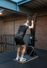 Inimene, kes kannab musta spordiriietust, kasutab siseruumides tellisseina taustal Concept2 SkiErg masinat.