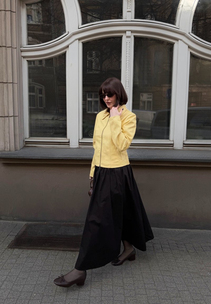 Femme en jupe noire, veste jaune et lunettes de soleil marchant sur le trottoir devant une grande fenêtre en arc avec du verre texturé.