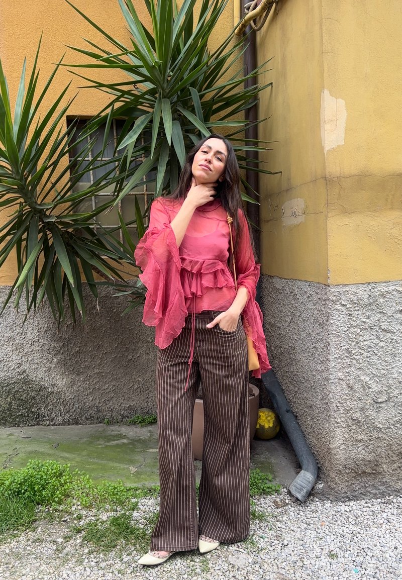 Femme en blouse rose transparente et pantalon marron à fines rayures, debout près d'un mur jaune et de plantes vertes, posant avec une main sur le cou et l'autre dans la poche.