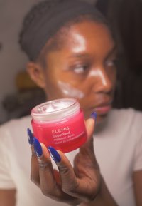 Vaaleanpunainen ELEMIS Superfood Midnight Facial -voiteen purkki, jonka sisällä valkoista voidetta ja läpinäkyvä kansi, pidetään sumennetun taustan edessä. Siniset kynnet näkyvissä.