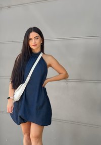 Woman with long dark hair wearing a sleeveless navy dress and a white crossbody bag, standing against a plain grey wall.