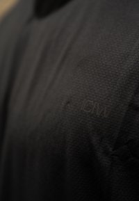 Μαύρο υφασμάτινο ύφασμα με διακριτό σχέδιο πλέγματος; διαθέτει ένα μικρό, ανάγλυφο λογότυπο στην επάνω αριστερή γωνία.