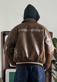 Veste aviateur en cuir marron avec poignets et ceinture côtelés, arborant un texte blanc "TURBO SPIRIT" au dos et une finition texturée.