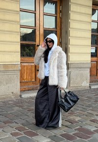 Béžová kožešinová bunda přes šedou mikinu, černé saténové kalhoty s širokým střihem a bílými pruhy, držící černou kabelku, stojící u dřevěných dveří.