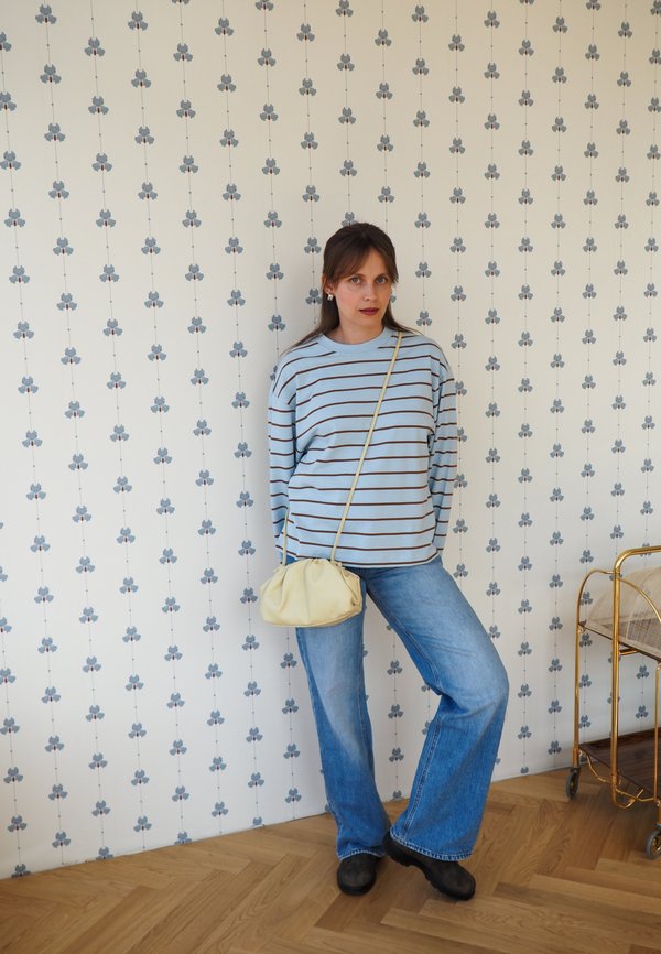 Mujer con jersey azul a rayas y vaqueros de pierna ancha está apoyada contra una pared con estampado sosteniendo un bolso beige, con un carrito dorado en el suelo de madera.