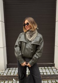 Veste surdimensionnée en similicuir vert olive avec larges revers, associée à une écharpe beige à motifs et un pantalon large foncé, tenant un sac fourre-tout noir tissé.
