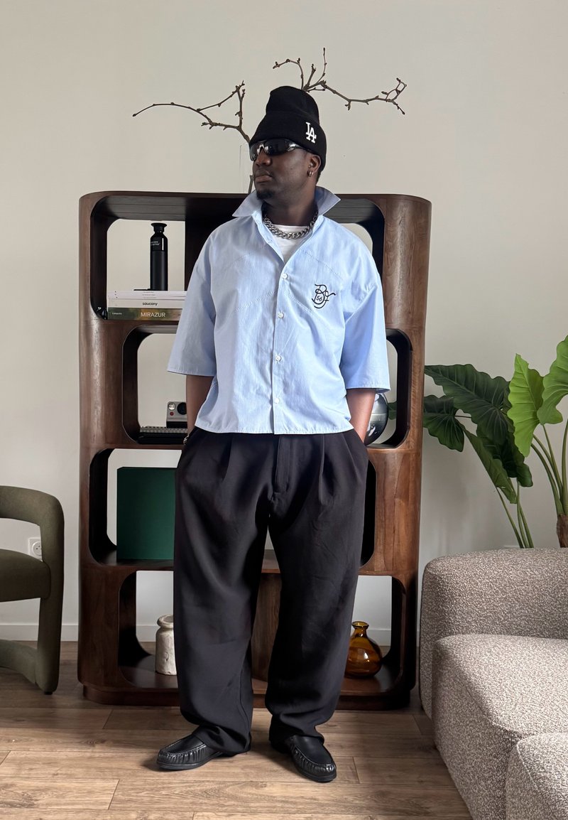 Homme portant une chemise bleu clair à manches courtes, un pantalon noir, des lunettes de soleil et un bonnet noir debout dans un salon moderne.