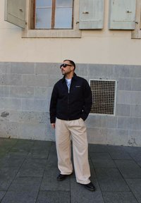 Schwarze Zip-Jacke, hellblauses gemustertes Hemd, beige weite Hose, schwarze Schuhe, Sonnenbrille. Graue Steinmauer mit Fenster im Hintergrund.