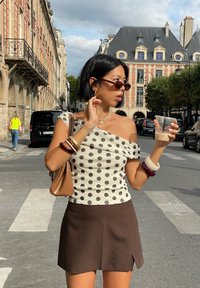 Top off-the-shoulder com bolinhas em creme e castanho, combinado com uma saia mini castanha. Acessórios incluem pulseiras em camadas e óculos de sol. A segurar uma bebida.