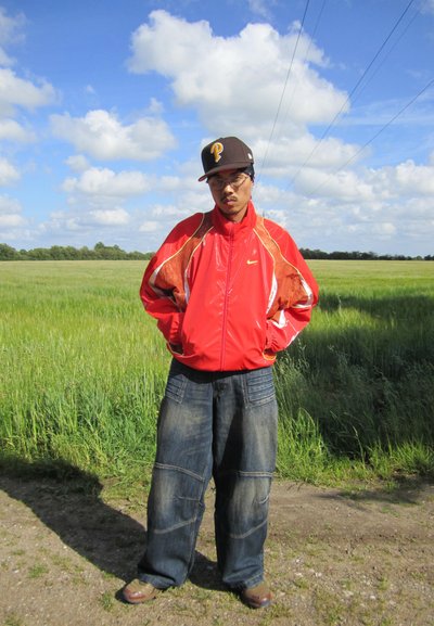 Chaqueta roja de Nike con acentos blancos, pantalones de mezclilla azul oversized, gorra marrón con logo amarillo, de pie en un camino de tierra con fondo de hierba verde.
