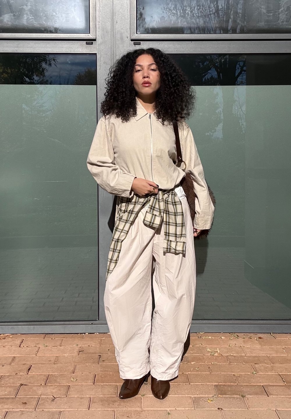 Woman with curly hair wearing a beige jacket and pants, a plaid shirt tied at her waist, brown boots, and carrying a brown shoulder bag, standing outdoors.