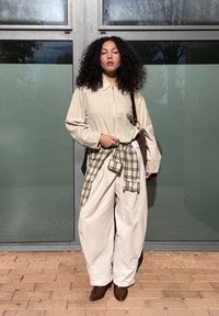 Woman with curly hair wearing a beige jacket and pants, a plaid shirt tied at her waist, brown boots, and carrying a brown shoulder bag, standing outdoors.
