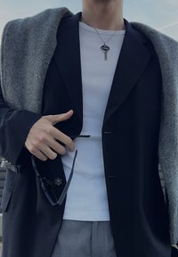 Personne portant un blazer noir sur une chemise blanche, un pantalon gris, un pull gris drapé sur les épaules, tenant des lunettes de soleil noires et un collier avec une clé.