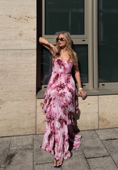 Mujer con vestido plisado con flores en tonos rosa y morado y gafas de sol, de pie junto a un muro de piedra al lado de una ventana grande bajo la luz del sol.