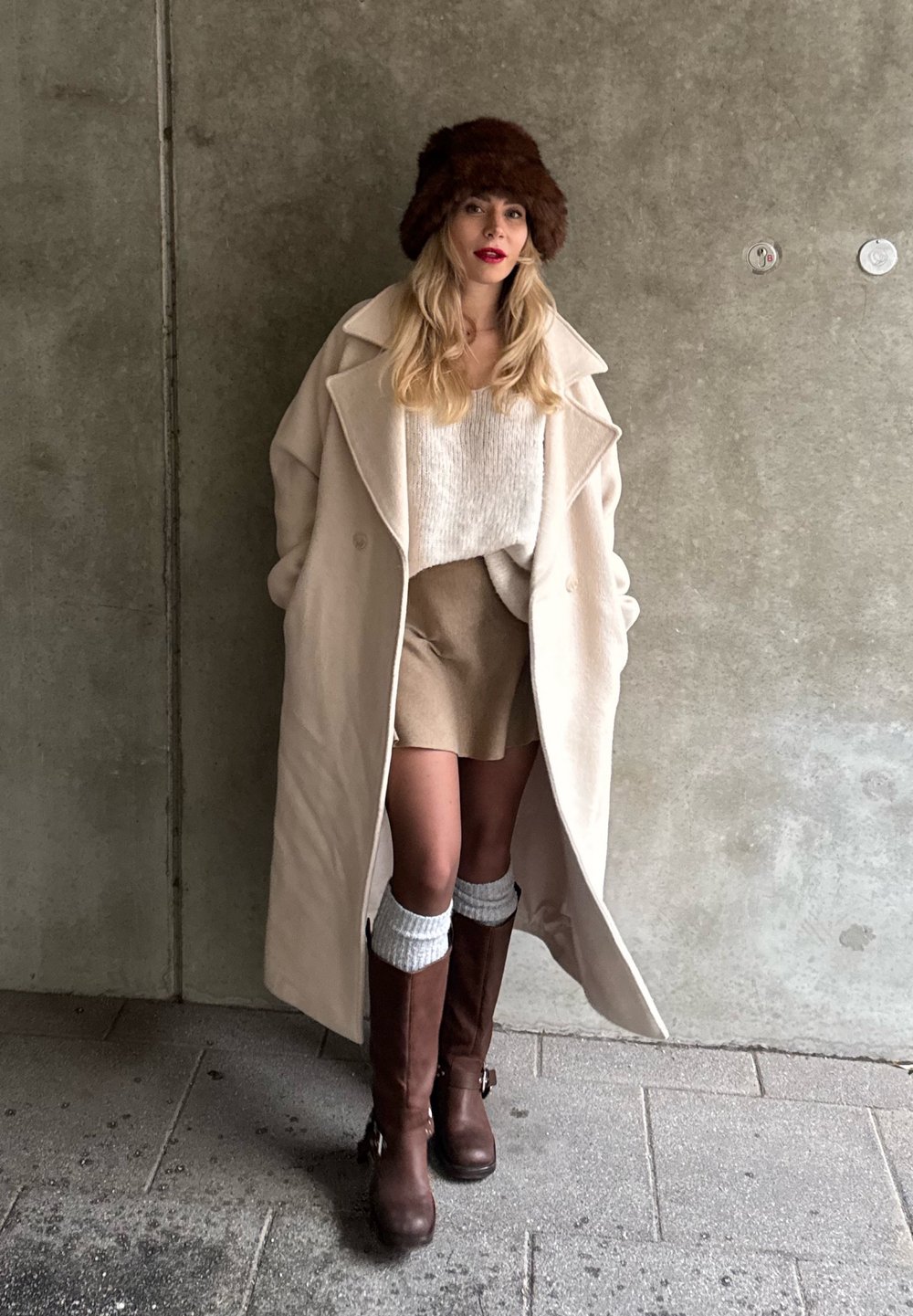Manteau surdimensionné beige, pull crème, jupe beige, bottes marron jusqu'au genou, chapeau en fourrure marron, avec des matériaux texturés et un look superposé.