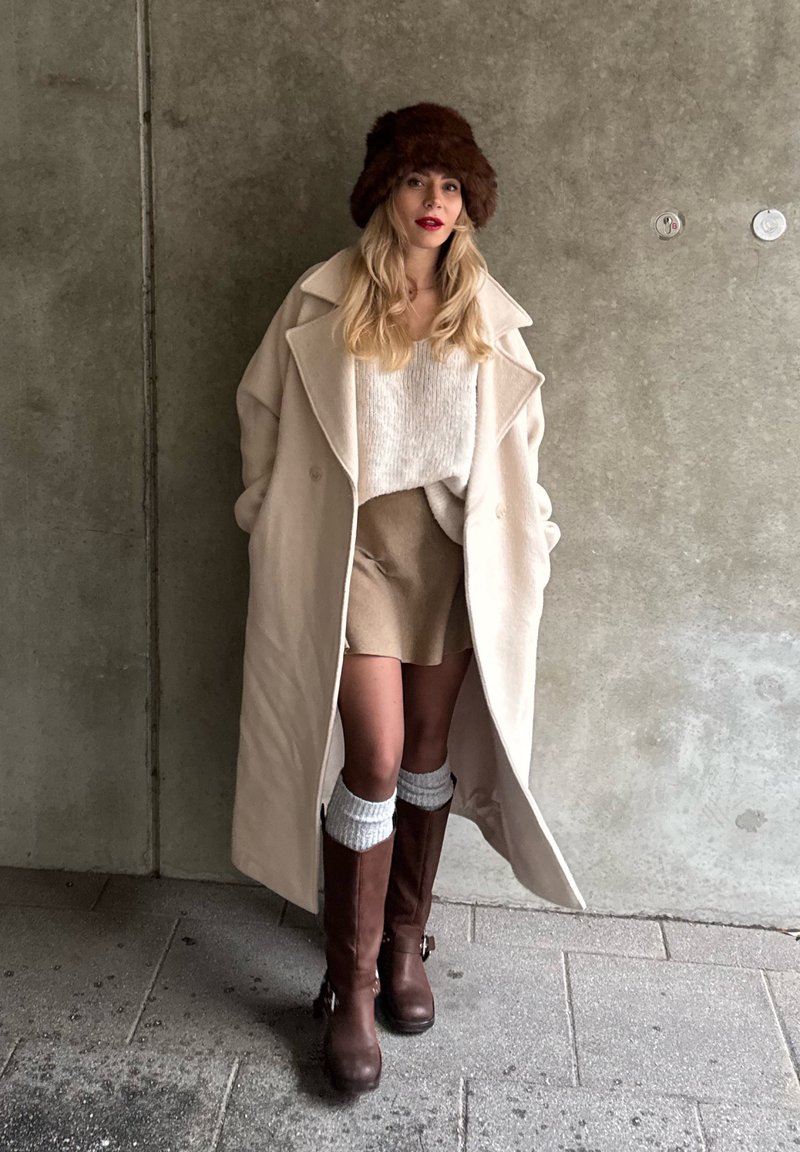 Manteau beige surdimensionné, pull crème, jupe beige, bottes marron montant jusqu'aux genoux, chapeau en fourrure marron, avec des matières texturées et un look en couches.