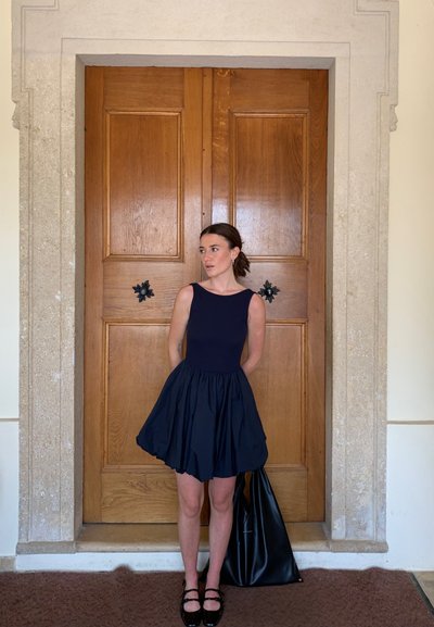 Vestido sin mangas de color azul marino con un torso ajustado y una falda acampanada, hecho de una tela suave. Bolso tote negro y sandalias. Puertas dobles de madera en el fondo.