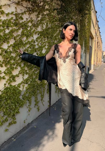 Mujer con peinado bob negro que lleva un vestido lencero crema con lunares y encaje negro, pantalones de cuero negro, sosteniendo una chaqueta de cuero negra, de pie junto a una pared cubierta de hiedra.