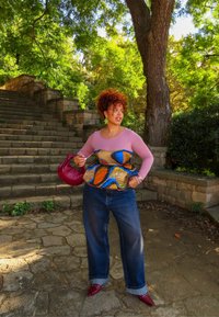 Kvinne med krøllete hår som har på seg en rosa topp, fargerik korsett, vide jeans og røde sko, står nær steintrappen i en grønn park.