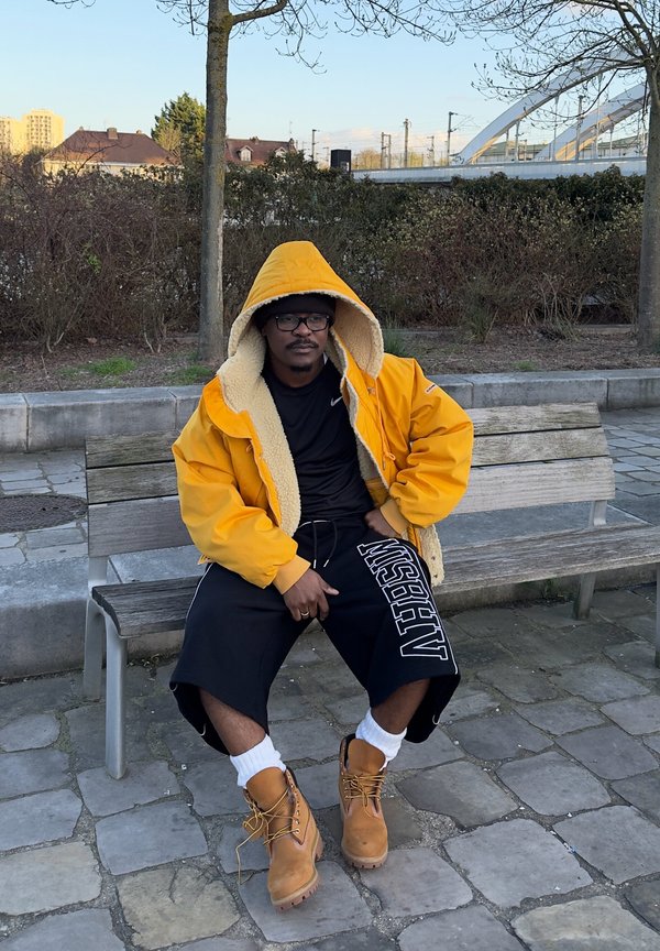 Man draagt een gele capuchonjas, zwarte korte broek en beige laarzen, zittend op een houten bankje in een stedelijke buitenruimte met bomen en een brug achter zich.