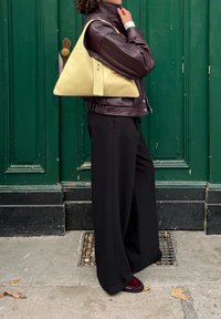 Driehoekige gele leren tas met een gladde textuur, vastgehouden naast een persoon in een donkerbruine leren jas en zwarte wijde broek.