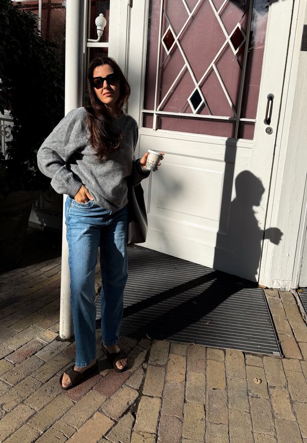 Mujer con gafas de sol, suéter gris y vaqueros, de pie sosteniendo una taza cerca de una puerta con paneles de vidrio decorativos bajo la luz del sol.
