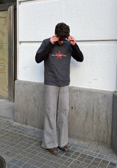 Joven ajustándose las gafas de sol, lleva una camisa negra de manga larga debajo de una camiseta gris con estrella roja y vaqueros grises de pierna ancha.