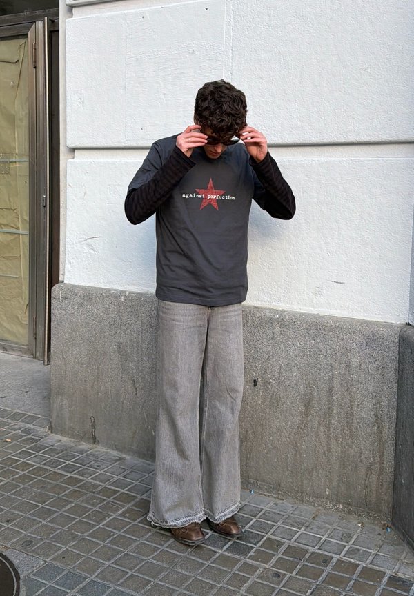 Jeune homme ajustant ses lunettes de soleil, portant un t-shirt noir à manches longues sous un t-shirt gris avec une étoile rouge et un jean gris à jambes larges.