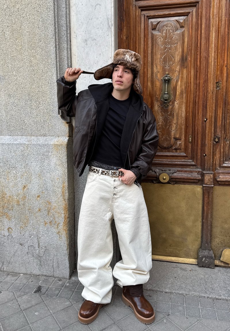 Jeune homme portant une veste en cuir marron, une chemise noire, un pantalon blanc ample, des bottes marron et un chapeau en fourrure, posant près d'une porte en bois ornée sur un trottoir en ville.