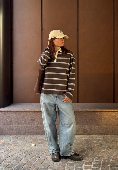 Mujer llevando una gorra beige, una camiseta de manga larga a rayas marrón, unos vaqueros anchos de color azul claro, zapatos marrones y una bolsa tote marrón.