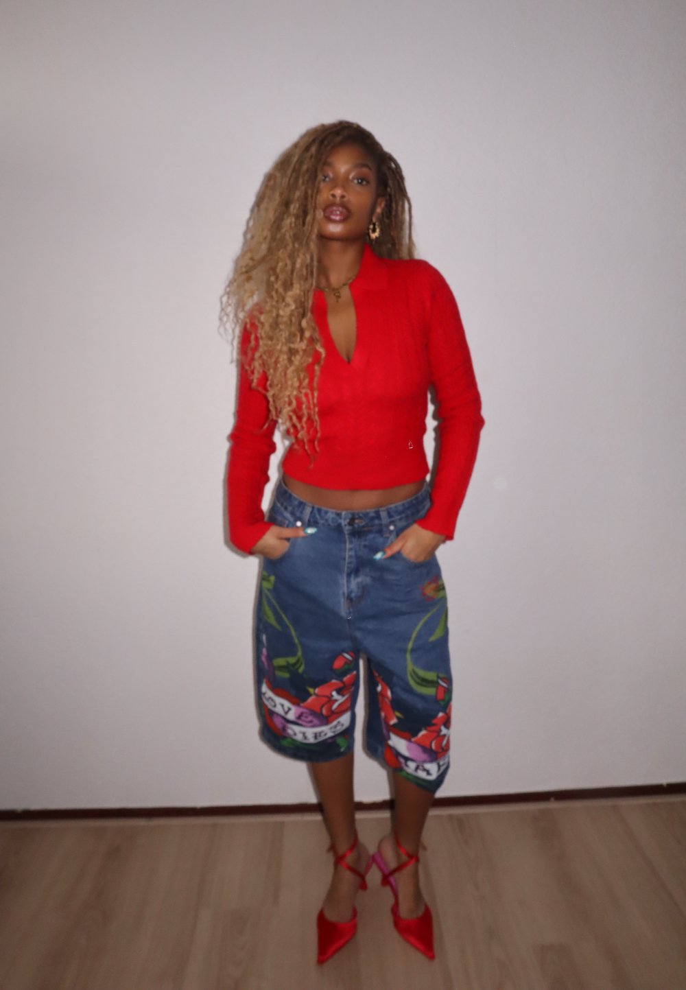 Rode ribbelige lange mouw top, denimshorts met bloemkunst en tekst, rode hoge hakken, textuurhaar, staand tegen een effen muur.