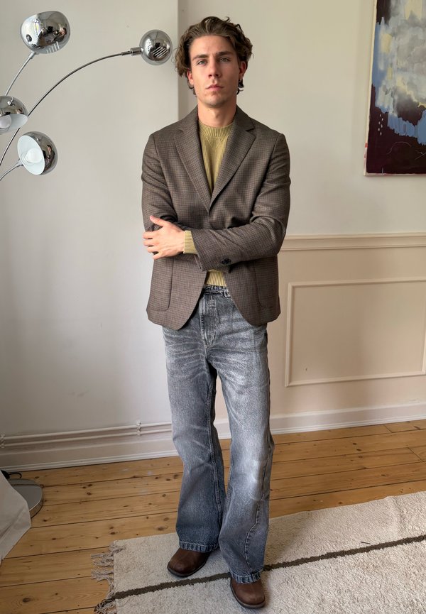 Young man with crossed arms wearing a brown blazer, mustard sweater, faded jeans, and brown shoes standing in a room with wooden floors and a lamp.